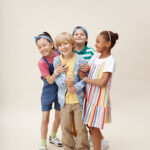 Vertical shot of multiethnic group of four little children in colorful clothes playing together while hugging excited boy wishing kid happy birthday against neutral background in studio, copy space